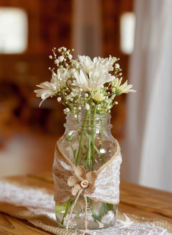 Vaso feito com frasco de vidro levando um tecido de juta com renda e botão de flor.