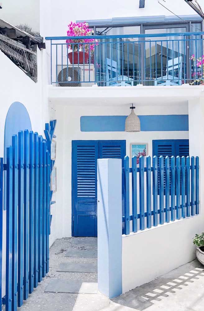 Sobrado simples e rústico com pintura branca, portão, grade e janelas de madeira na cor azul.