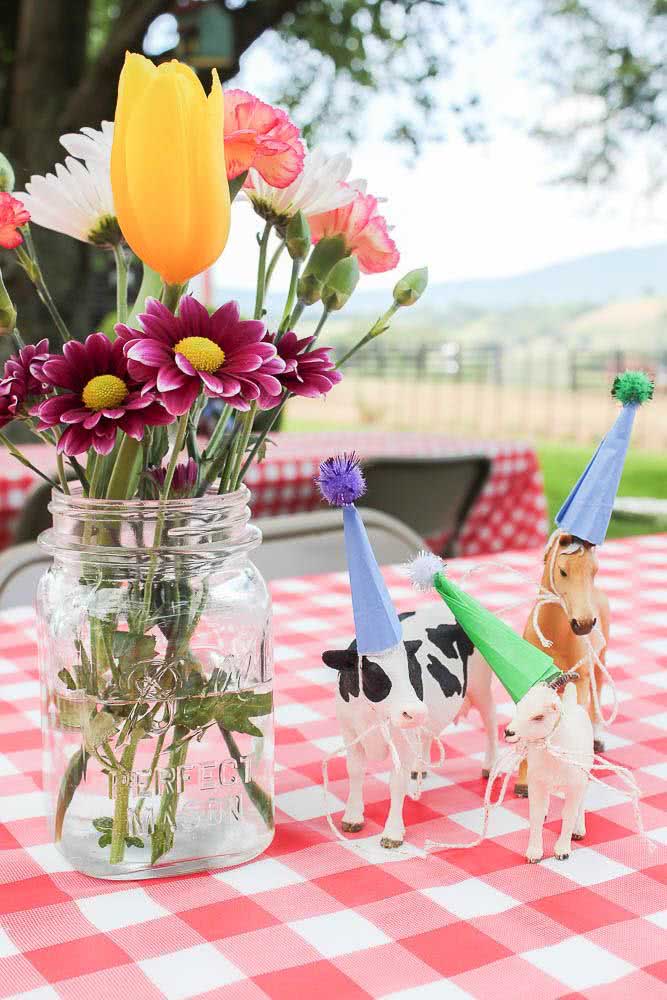 Mesa com toalha quadriculada, um belíssimo vaso de flores e pequenos animais com chapéuzinhos.