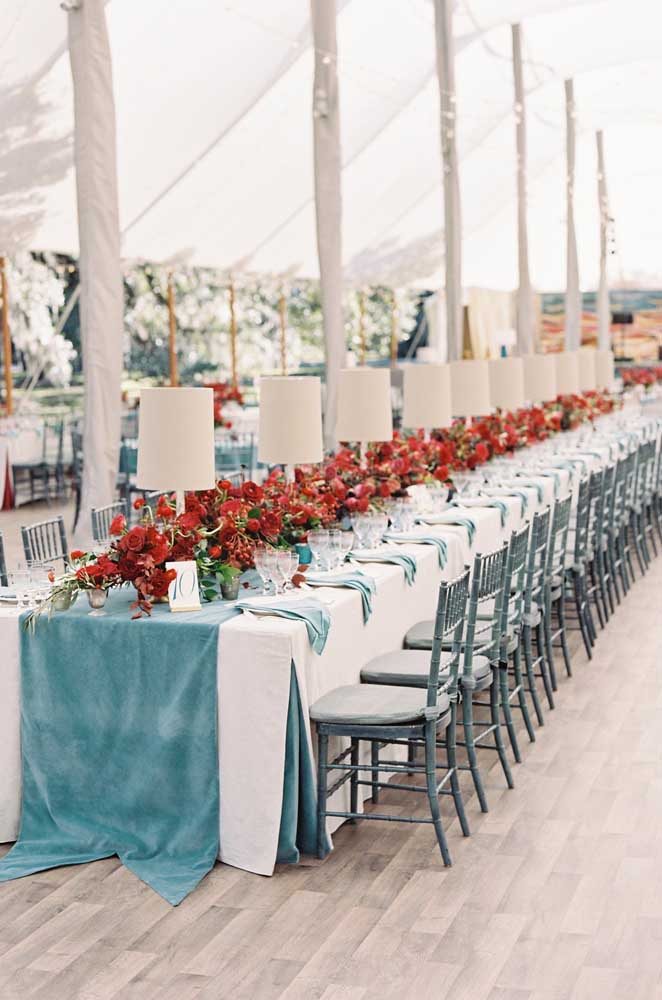 Mesa de casamento com toalha, guardanapos e cadeiras azuis. Os arranjos de flores centrais foram elaborados com flores vermelhas.