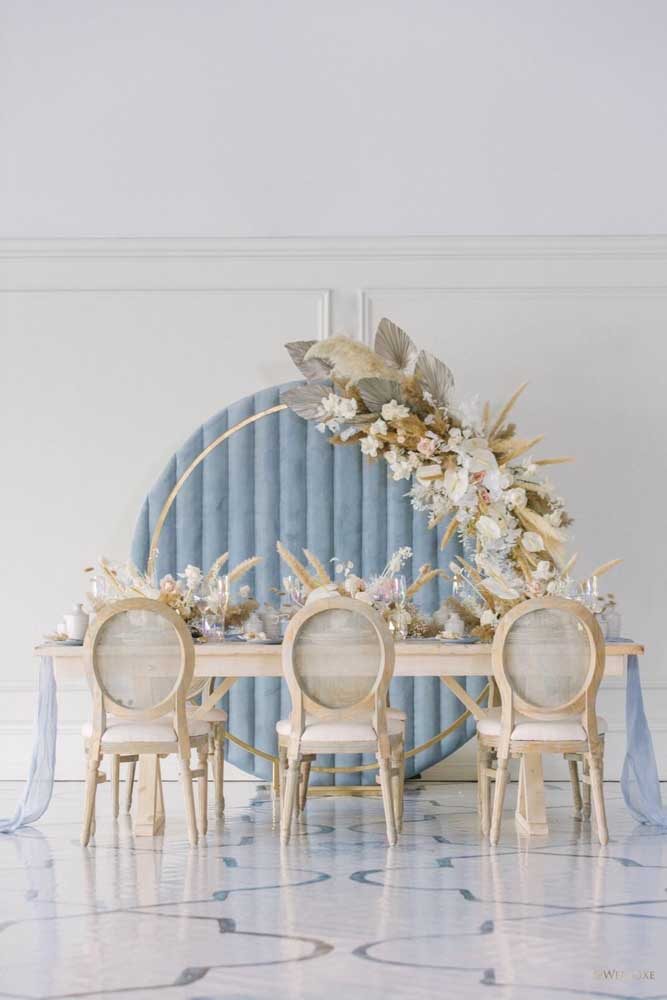 Painel para mesa de bolo de casamento azul com detalhes em dourado e arranjos de flores.
