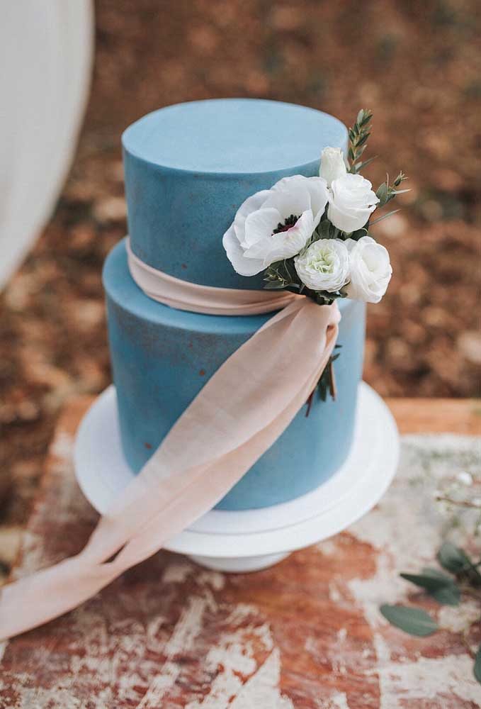 Bolo fake azul de casamento com buquê de flores brancas e fita de tecido claro.