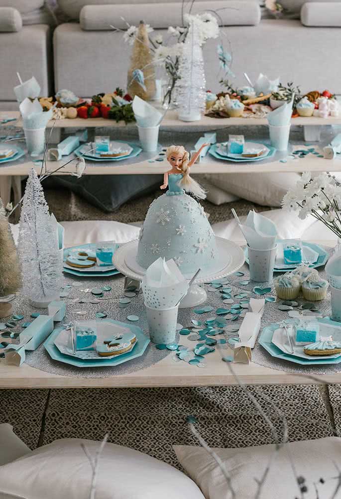 Decoração de festa Frozen em mesa baixa no chão com pratos e bolo personalizado com a boneca do tema.