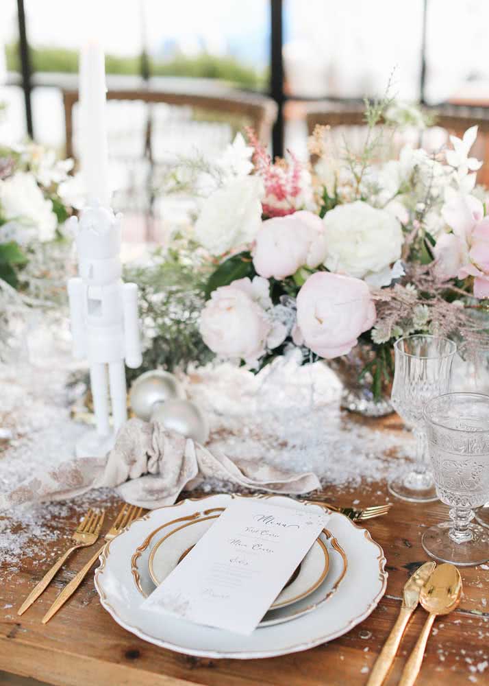 Mesa de Natal com flores brancas e claras e pratos com talheres dourados.