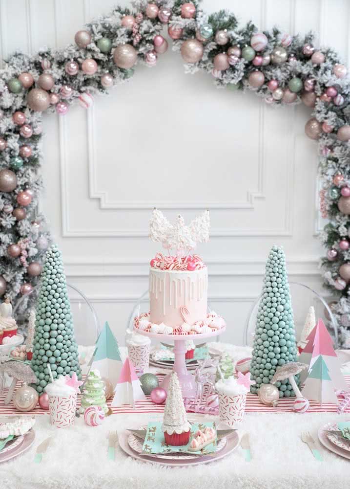 Mesa delicada com arco de bolas na cor rosa e bolo de Natal no centro da mesa.