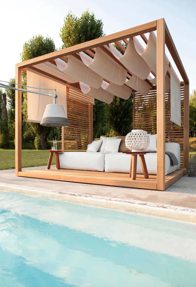 Área da piscina com estrutura de madeira para abrigar sofá grande com pergolado e cobertura de tecido.