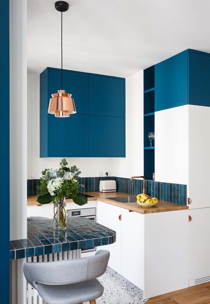 Cozinha em L branca pequena e compacta com bancada de madeira e armários superiores na cor azul marinho.