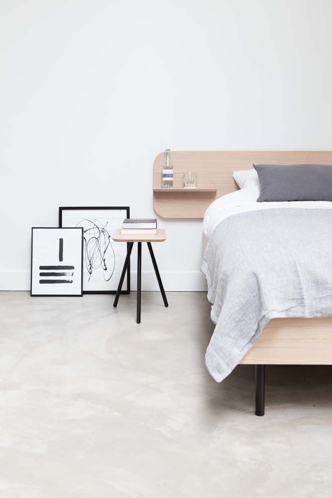 Quarto de casal minimalista com cama e cabeceira na madeira clara e quadros apoiados no chão.