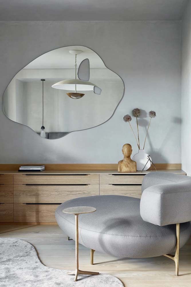 Modelo de espelho com formato curvo diferenciado em sala de estar minimalista com tons de cinza e móveis de madeira claro.