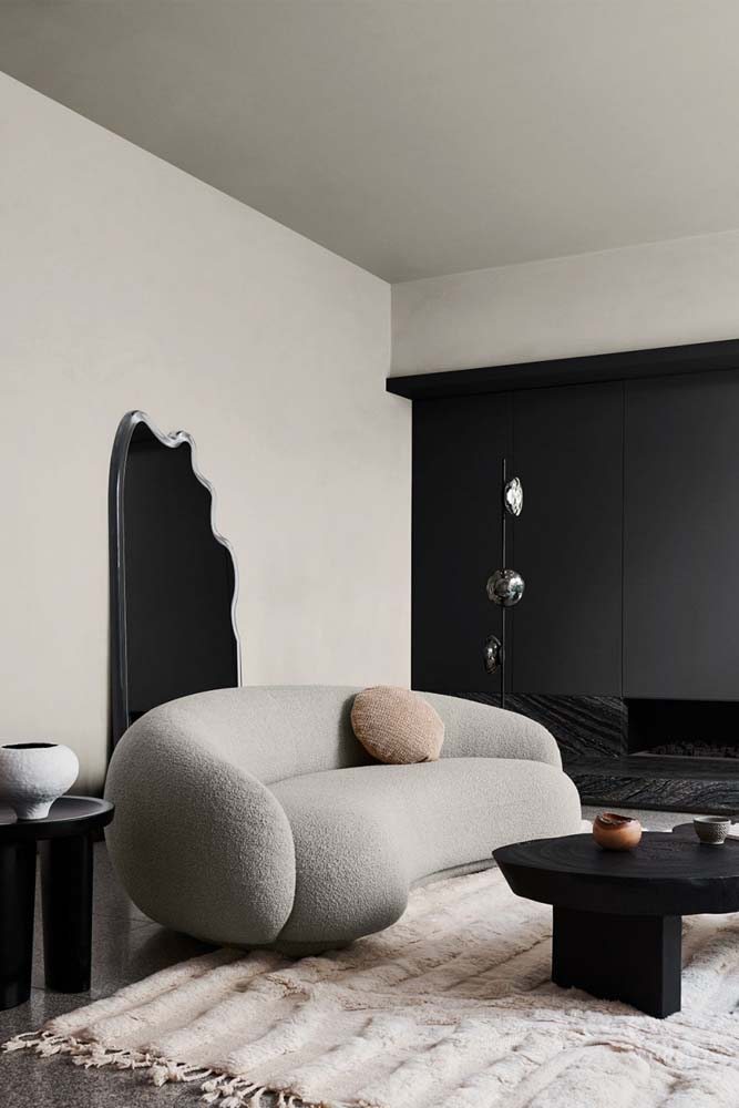 Sala moderna e minimalista com sofá curvo, mesa de centro e lateral na cor preta e espelho decorativo atrás do sofá apoiado na parede.