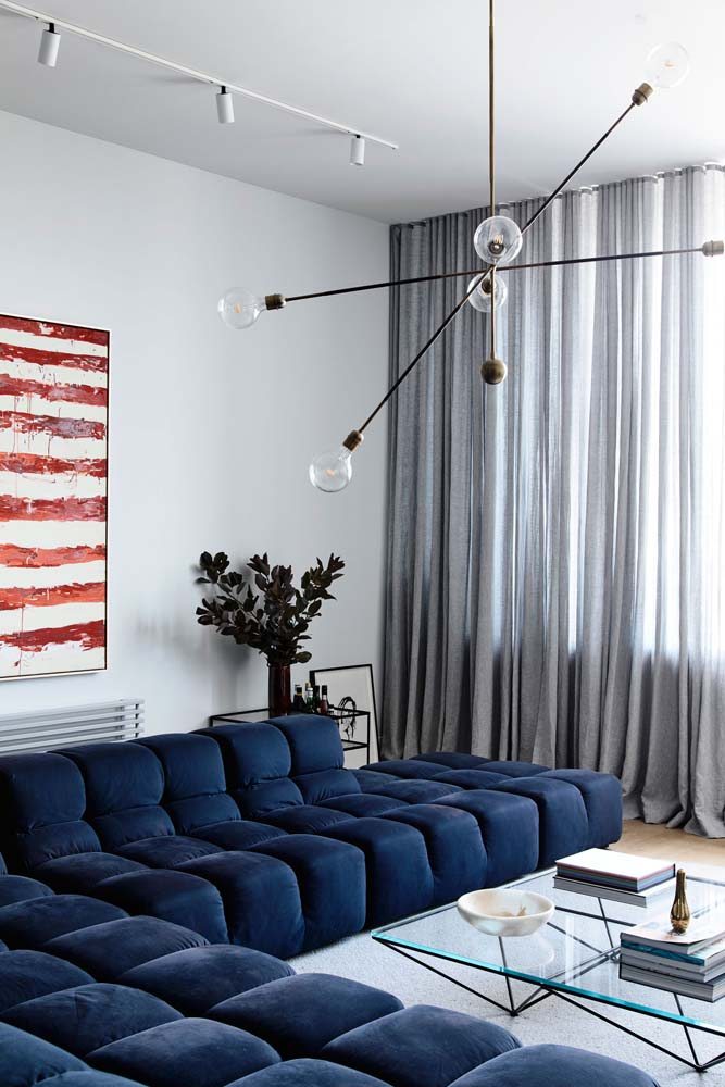 Sala com pintura branca, cortina na cor cinza e sofá de canto com tecido azul escuro.