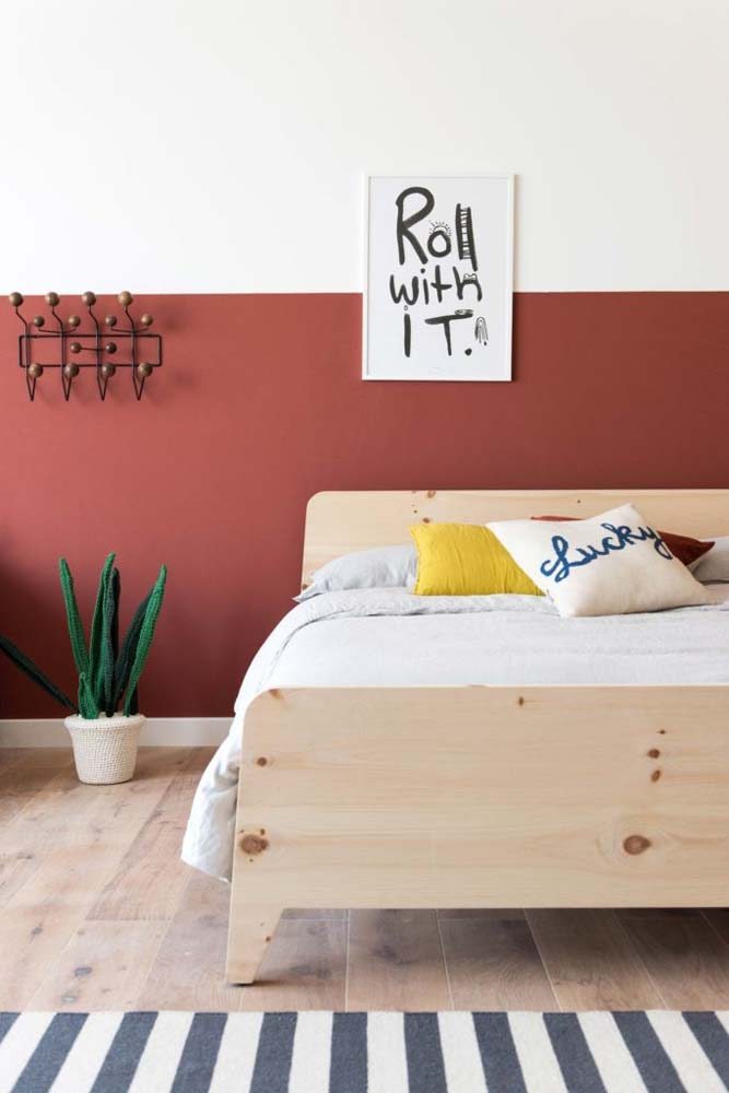 Quarto minimalista com meia parede pintada em tons terrosos e cama de madeira clara.