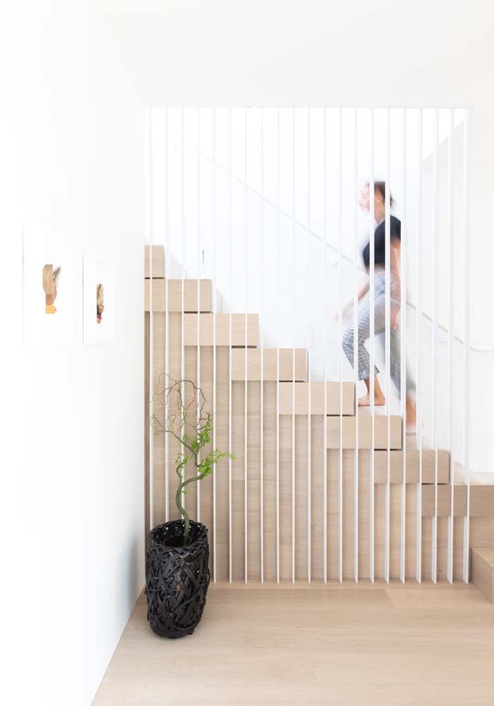 Escada de madeira clara com proteção vazada lateral em tiras metálicas com pintura branca.
