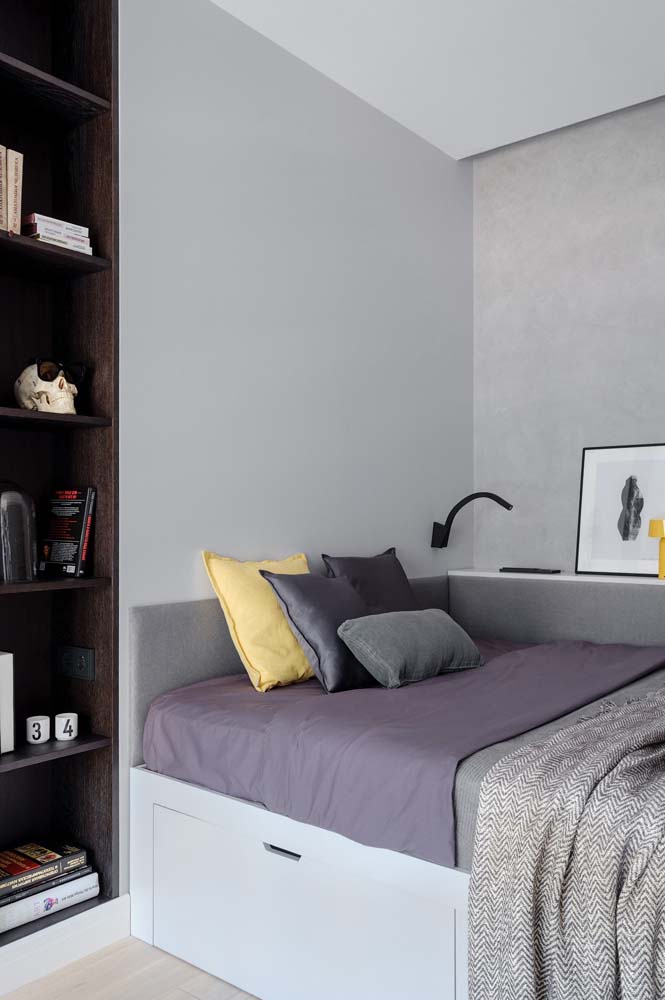 Cantinho do quarto com pintura cinza, cama compacta com cabeceira estofada e prateleira de madeira escura na lateral.