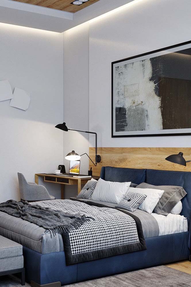 Modelo de quarto de solteiro com escrivaninha, quadro grande, cabeceira de madeira clara e cama azul marinho.