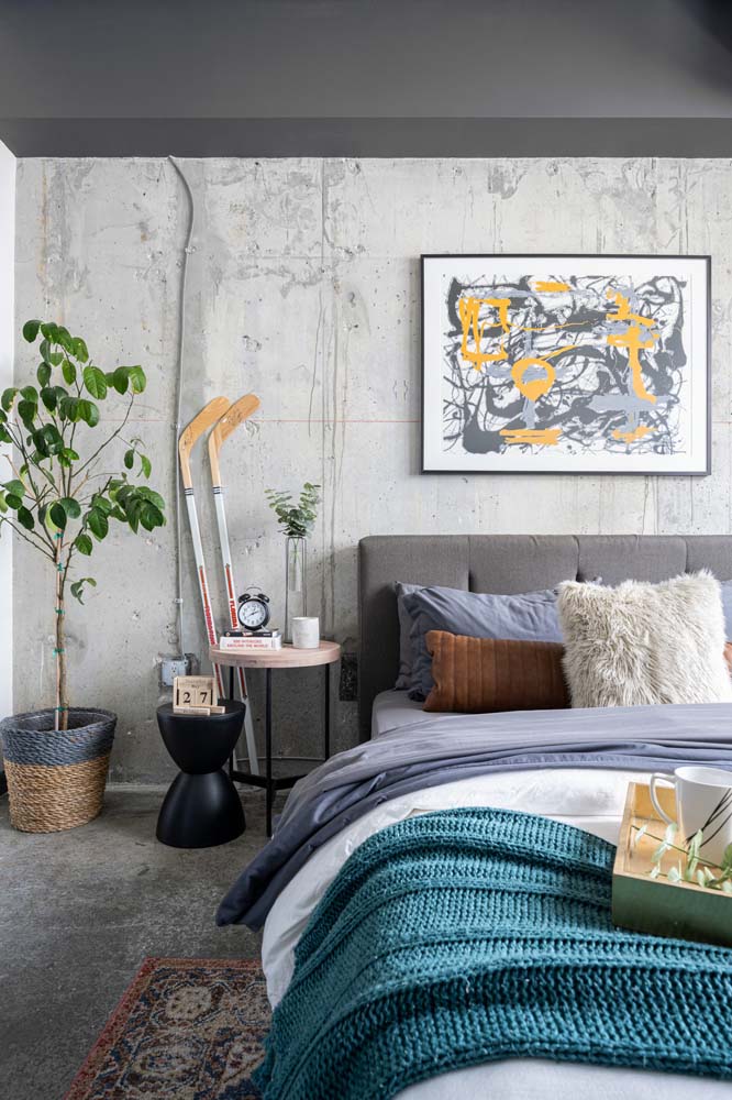 Quarto grande com concreto aparente nas paredes, piso de cimento queimado e um toque de cor na roupa de cama, quadros e vaso de plantas.